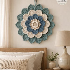 May include: A circular wall hanging featuring a layered, multi-petaled flower design crafted from crochet yarn. The piece displays concentric rings of petals in shades of teal, cream, and blue, with a small loop at the top for hanging.