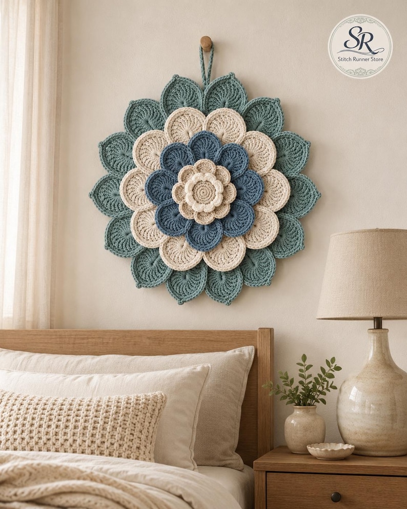 May include: A circular wall hanging featuring a layered, multi-petaled flower design crafted from crochet yarn. The piece displays concentric rings of petals in shades of teal, cream, and blue, with a small loop at the top for hanging.