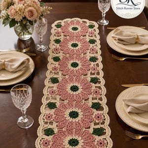 May include: A rectangular crochet table runner featuring a repeating pattern of large dusty pink flowers with dark green centers, accented by smaller pink flowers and dark green leaves, all bordered by an off-white scalloped lace edge.
