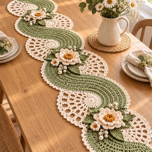 Puede incluir: Un camino de mesa de ganchillo largo y sinuoso que presenta un patrón alterno de motivos de encaje circulares de color crema y secciones onduladas de color verde salvia. El camino está adornado con flores tridimensionales color crema con centros amarillos y hojas verdes.