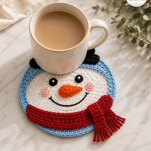 Peut inclure: Un dessous de verre au crochet en forme de bonhomme de neige, avec un visage blanc, des yeux noirs, un nez orange, des joues roses et un sourire noir. Le bonhomme de neige porte un bonnet bleu clair et une écharpe rouge texturée à franges.