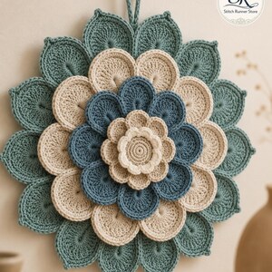May include: A circular crochet wall hanging featuring a layered flower design in shades of teal, cream, and blue. The piece has a central cream flower, surrounded by concentric rings of blue and cream petals, finished with a large teal outer layer and a hanging loop at the top. The logo SR Stitch Runner Store is visible in the corner.