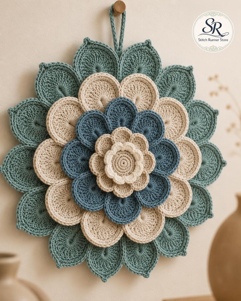 May include: A circular crochet wall hanging featuring a layered flower design in shades of teal, cream, and blue. The piece has a central cream flower, surrounded by concentric rings of blue and cream petals, finished with a large teal outer layer and a hanging loop at the top. The logo SR Stitch Runner Store is visible in the corner.