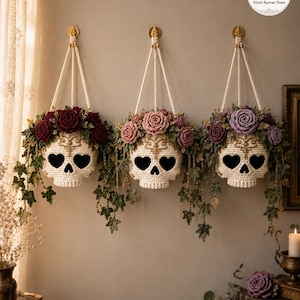 Puede incluir: Tres maceteros colgantes de calavera tejidos a ganchillo en color blanco roto, adornados con una corona de flores tejidas y enredaderas verdes colgantes. Las calaveras tienen cuencas oculares en forma de corazón y detalles dorados.