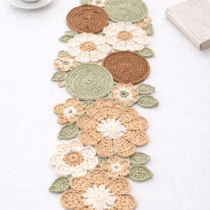 May include: A handmade crochet table runner with a floral design. The runner is made of beige, brown, and green yarn, with flower and leaf shapes. A cup of coffee and a book are in the background.