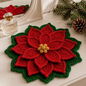 May include: A round, crocheted poinsettia-shaped mat featuring layered red petals, a dark green leaf-shaped border, and a central cluster of small, textured yellow spheres. The design mimics a blooming flower with intricate yarn stitching.