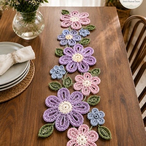 May include: A crochet table runner featuring a floral pattern with large and small flowers in shades of lavender, light pink, and periwinkle blue, accented by green crocheted leaves. The design includes a circular logo reading SR Stitch Runner Store.