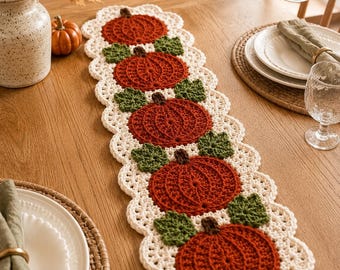 May include: A rectangular crochet table runner featuring five circular orange pumpkins with brown stems and green leaves, bordered by a cream-colored scalloped edge. The runner is centered on a wooden surface, showcasing intricate yarn textures.