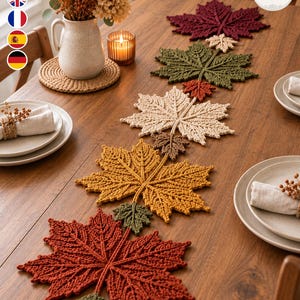 May include: A crochet table runner featuring a series of interconnected maple leaves in autumn colours including deep red, olive green, cream, mustard yellow, and brown, arranged in a linear pattern down the centre of a wooden dining table.