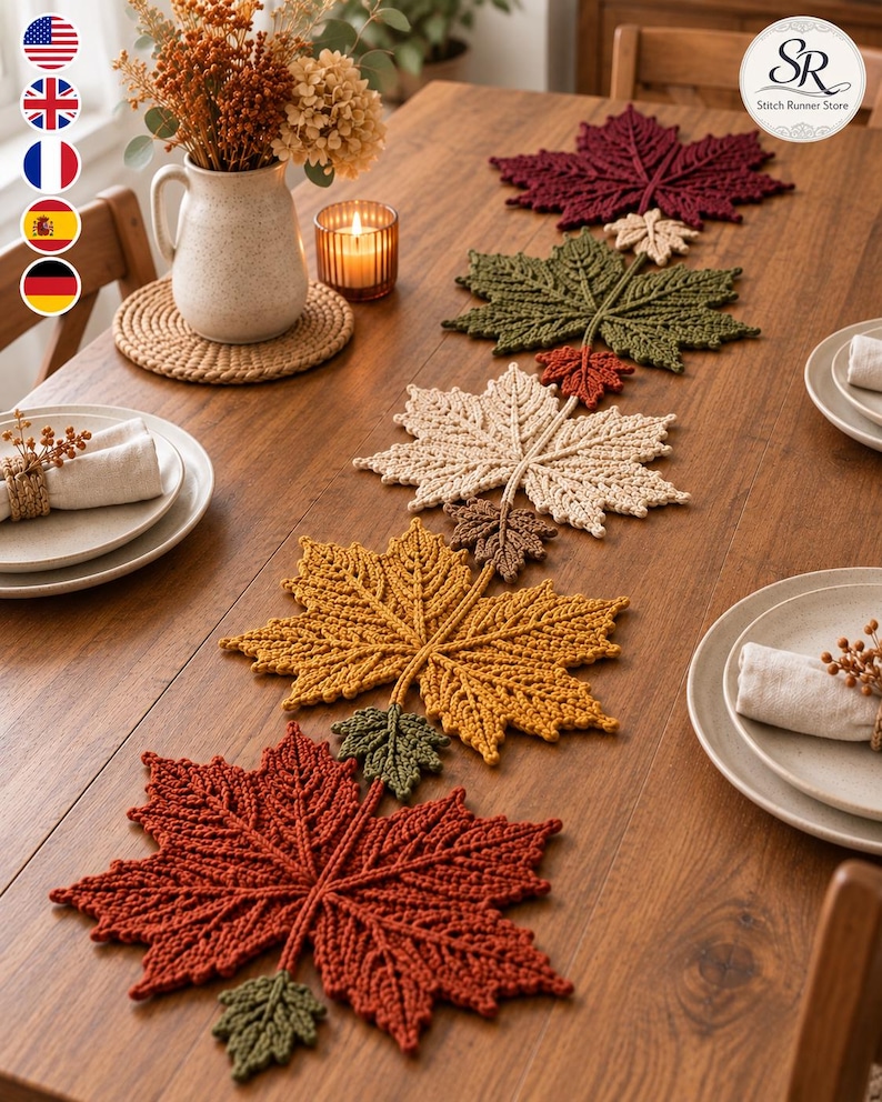 May include: A crochet table runner featuring a series of interconnected maple leaves in autumn colours including deep red, olive green, cream, mustard yellow, and brown, arranged in a linear pattern down the centre of a wooden dining table.