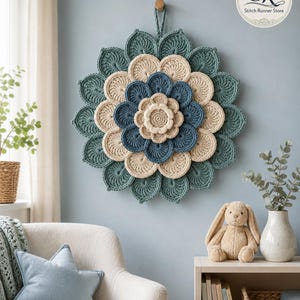May include: A circular wall hanging featuring a layered, multi-colored crochet flower design. The piece consists of concentric rings of petals in shades of cream, dark blue, and muted teal, with a small loop at the top for hanging on a wall.