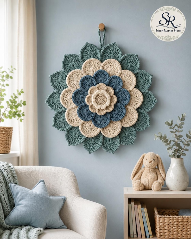 May include: A circular wall hanging featuring a layered, multi-colored crochet flower design. The piece consists of concentric rings of petals in shades of cream, dark blue, and muted teal, with a small loop at the top for hanging on a wall.
