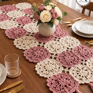 May include: A handmade crochet table runner in alternating dusty rose and cream circular patterns. A vase of roses sits atop the runner, with gold cutlery and a glass of water on the wooden table. Tassels hang from the end of the runner.