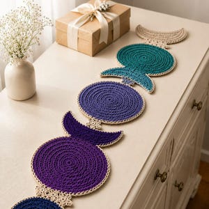 Puede incluir: Caminos de mesa de ganchillo hechos a mano en tonos azules, turquesa y morado, con ribete color crema. Los caminos de mesa están colocados sobre una superficie blanca. En el fondo hay una caja de regalo y un jarrón con flores blancas.