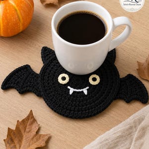 May include: A black crocheted bat coaster featuring two cream-colored circular eyes and white stitched fangs. The coaster is circular with two pointed ears at the top and two extended wings on the sides, supporting a white ceramic mug filled with black coffee.