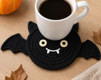 May include: A black crocheted bat coaster featuring two cream-colored circular eyes and white stitched fangs. The coaster is circular with two pointed ears at the top and two extended wings on the sides, supporting a white ceramic mug filled with black coffee.