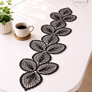 May include: A black, leaf-shaped lace table runner is displayed on a white table. The runner is made of delicate, open-weave lace and is arranged in a flowing pattern. A cup of coffee and a small potted plant are also on the table.