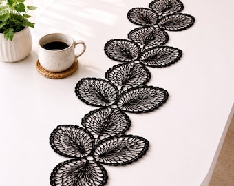 May include: A black, leaf-shaped lace table runner is displayed on a white table. The runner is made of delicate, open-weave lace and is arranged in a flowing pattern. A cup of coffee and a small potted plant are also on the table.