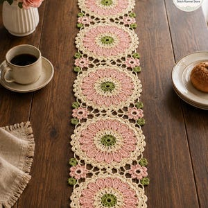 May include: A long, rectangular crochet table runner featuring a repeating pattern of large, circular floral motifs in shades of cream, soft pink, and olive green. The design includes smaller pink flower accents between the main circles.