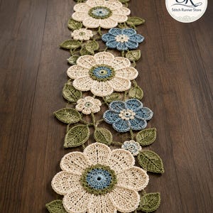 May include: A long, rectangular crochet table runner featuring a floral pattern with large cream-coloured flowers, smaller blue flowers, and green leaves. The design includes intricate stitching and a circular logo in the corner reading SR Stitch Runner Store.