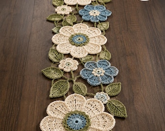 May include: A long, rectangular crochet table runner featuring a floral pattern with large cream-colored flowers, smaller blue flowers, and green leaves. The design includes intricate stitching and a circular logo in the corner reading SR Stitch Runner Store.