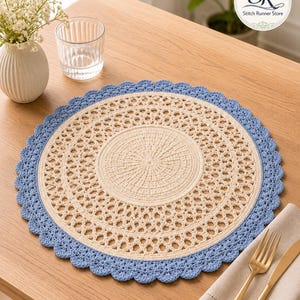 May include: A round, cream-colored crochet placemat with a light blue scalloped edge. The placemat has an openwork design and is placed on a wooden table. A glass of water, a vase with flowers, and silverware are also on the table.