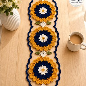 Puede incluir: Un camino de mesa rectangular de ganchillo con tres grandes motivos florales circulares en tonos amarillo mostaza, azul marino y blanco. El camino tiene un borde festoneado en azul marino y pequeños detalles de hojas verdes.