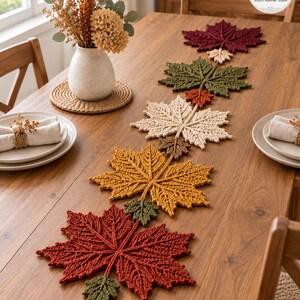 May include: A crochet table runner featuring a series of interconnected maple leaves in autumn colours including deep red, olive green, cream, and golden yellow, arranged in a linear pattern down the centre of a wooden dining table.