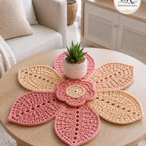 Può includere: Un centrotavola a forma di fiore all'uncinetto nei toni del rosa e del beige, con una piccola pianta grassa in vaso al centro. Il centrotavola è su un tavolino rotondo in legno. Il testo "COFFEE TABLE" è in basso.