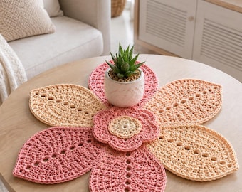 May include: A crocheted flower-shaped table centerpiece in shades of pink and beige, with a small potted succulent in the center. The centerpiece is on a round wooden coffee table. The text "COFFEE TABLE" is at the bottom.