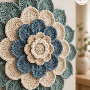 May include: A circular crochet wall hanging featuring layered flower petals in shades of sage green, cream, and dusty blue. The design consists of concentric rings of stitched petals radiating from a central cream-colored flower center.