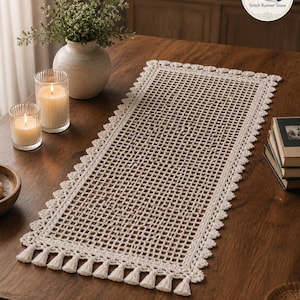 Puede incluir: Camino de mesa rectangular de ganchillo color crema con un diseño de enrejado y un borde festoneado. El camino de mesa está adornado con pequeñas borlas a lo largo del borde inferior. Se muestra sobre una mesa de madera oscura, con velas y un jarrón.