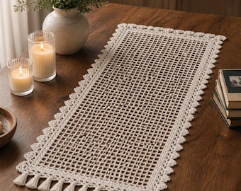 May include: A cream-colored, rectangular crochet table runner with a lattice design and a scalloped edge. The runner is adorned with small tassels along the bottom edge. The runner is displayed on a dark wooden table, alongside candles and a vase.