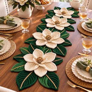 Può includere: Un lungo runner da tavola decorativo caratterizzato da quattro grandi fiori di magnolia realizzati all'uncinetto in tre dimensioni, con petali color crema e centri strutturati, circondati da foglie verde scuro con accenti in filo dorato.