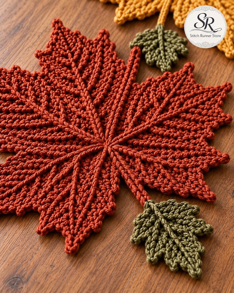 May include: A large, rust-coloured crocheted maple leaf with prominent textured veins, accompanied by two smaller olive green crocheted leaves attached by stems. A circular logo in the corner displays the text SR Stitch Runner Store.