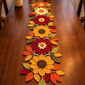 May include: A long, decorative crochet table runner featuring a series of interconnected flowers and leaves in autumn colours including deep red, bright yellow, orange, cream, and various shades of green, arranged in a vertical, symmetrical pattern.