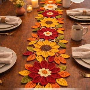 Puede incluir: Camino de mesa de ganchillo con diseño floral. El camino de mesa presenta flores rojas, amarillas y naranjas y hojas verdes. El texto "Styled for Your Table" se muestra en la parte inferior. El camino de mesa está colocado sobre una mesa de madera preparada para una comida.