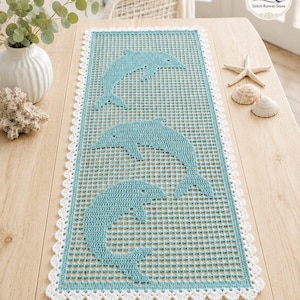 May include: A rectangular, turquoise table runner with three dolphin designs. The dolphins are a lighter shade of turquoise. The runner has a white, scalloped lace border. The runner is displayed on a light wood table.