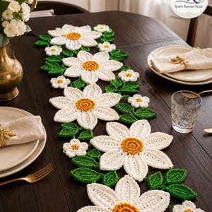 K&ouml;nnte beinhalten: Ein geh&auml;kelter Tischl&auml;ufer mit gro&szlig;en wei&szlig;en G&auml;nsebl&uuml;mchenmotiven, gelben Bl&uuml;tenmitten und gr&uuml;nen Bl&auml;ttern. Das florale Design ist in einem durchgehenden Muster entlang des L&auml;ufers angeordnet und zeigt detaillierte Maschenstrukturen.