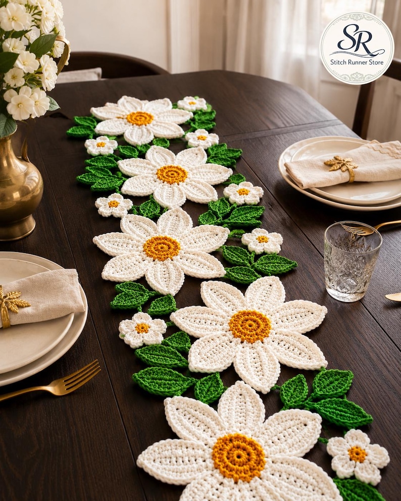 K&ouml;nnte beinhalten: Ein geh&auml;kelter Tischl&auml;ufer mit gro&szlig;en wei&szlig;en G&auml;nsebl&uuml;mchenmotiven, gelben Bl&uuml;tenmitten und gr&uuml;nen Bl&auml;ttern. Das florale Design ist in einem durchgehenden Muster entlang des L&auml;ufers angeordnet und zeigt detaillierte Maschenstrukturen.