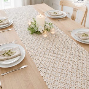 Op de afbeelding: Crèmekleurige tafelloper met open weefsel en geschulpte rand, op een lichte houten tafel. De loper is versierd met een grote witte kaars, kleine votiefkaarsen en bloemaccenten. Eetplaatsen met witte borden en linnen servetten zijn zichtbaar.