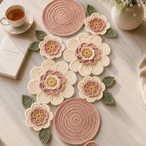 Könnte beinhalten: Handgefertigte Häkelartikel, darunter blumenförmige Untersetzer und runde Tischsets in Rosa, Creme und Gelb. Grüne Blätter und eine Teetasse ergänzen das Arrangement und erzeugen eine gemütliche, handwerkliche Atmosphäre. Maße in cm.