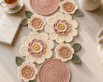 Könnte beinhalten: Handgefertigte Häkelartikel, darunter blumenförmige Untersetzer und runde Tischsets in Rosa, Creme und Gelb. Grüne Blätter und eine Teetasse ergänzen das Arrangement und erzeugen eine gemütliche, handwerkliche Atmosphäre. Maße in cm.