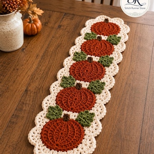 Puede incluir: Un camino de mesa de ganchillo rectangular con cinco calabazas circulares de color naranja óxido con tallos marrones y hojas verdes, bordeado por un borde festoneado de color crema. El diseño es repetitivo y centrado con un logo en la esquina.