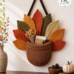 May include: A wall-hanging crochet basket in brown, featuring a fan of textured leaves in shades of orange, yellow, olive green, and tan. The basket is suspended by a dark brown strap and contains dried flowers and cards. Logo reads Stitch Runner Store.