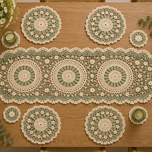 May include: A crochet table runner featuring three large circular motifs in cream and sage green, surrounded by a delicate floral lace border. The set includes four matching circular placemats and four small coasters in the same colour scheme.