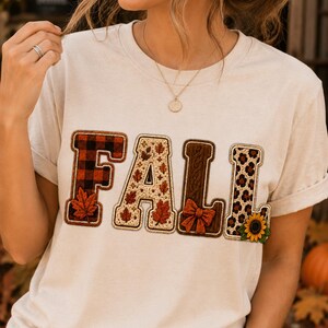 May include: A cream-colored short-sleeve t-shirt featuring the word FALL in large, textured letters. The F has a red and black buffalo plaid pattern with a leaf, the A has a leaf print, the L has a bow, and the second L has leopard print.