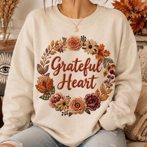 Op de afbeelding: Een crèmekleurige sweater met ronde hals en een centrale afbeelding van een bloemenkrans in herfsttinten zoals oranje, bruin en bordeaux. De tekst "Grateful Heart" staat in een donkerbruin cursief lettertype in het midden van de krans.