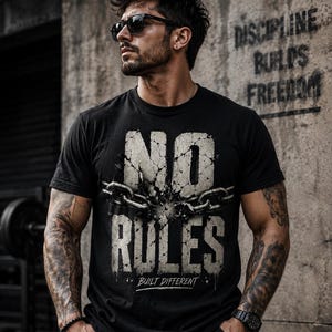 May include: A black short-sleeved t-shirt featuring a graphic print with the text NO RULES in a distressed, cracked stone texture. A metallic chain is depicted breaking across the center of the text, with the words BUILT DIFFERENT printed below.