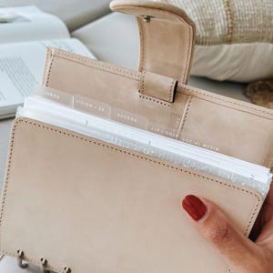 Op de afbeelding: Een beige lederen persoonlijke planner met een drukknoopsluiting en metalen ringband. De binnenzijde bevat doorschijnende tabbladen met teksten zoals INBOX, VISION / 23, AGENDA, L+P / ODSP en SOCIAL MEDIA, met witte papieren pagina's.